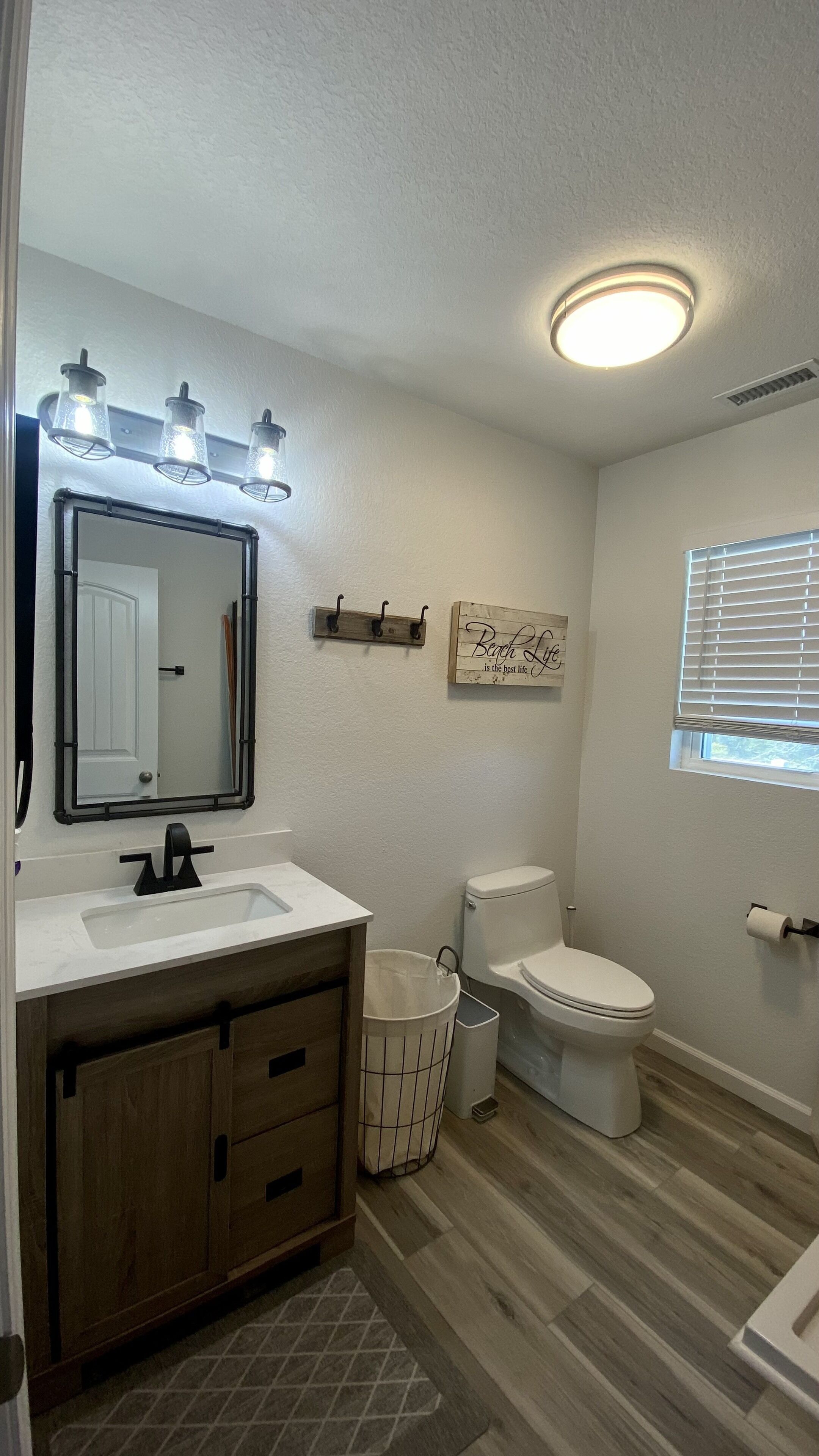 A bathroom with a white sink, wooden cabinets, a toilet, a trash can, and a window with blinds.