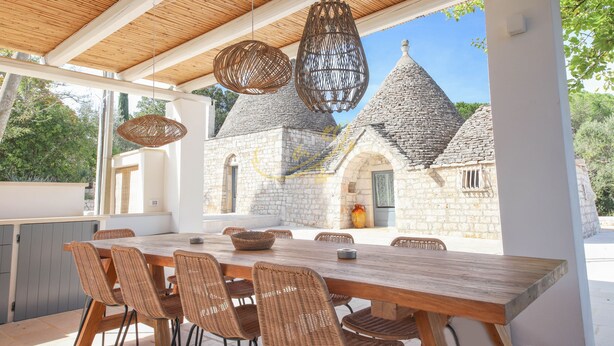 Un espace extérieur aménagé pour les repas, avec une table en bois et des chaises en osier, donnant sur un bâtiment en pierre surmonté de toits coniques.
