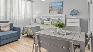 A modern bedroom with a bed, a blue sofa, a dining table with chairs, a ceiling fan, and a painting of a beach scene. - Beachfront Cozy Loft  Walk 2 Eat Isla Verde Beach (Carolina)