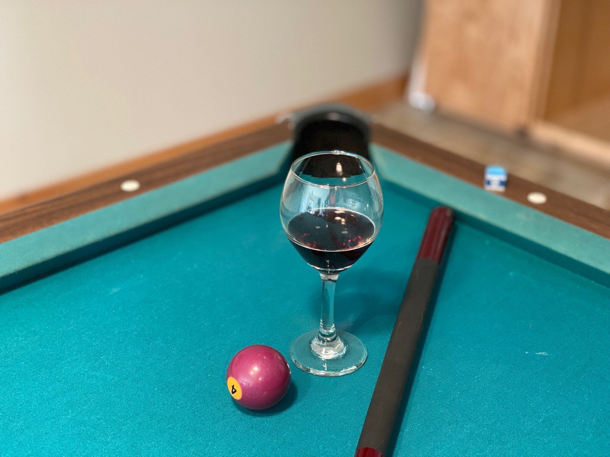 A pool table with a glass of red wine, a cue stick, and a billiard ball.