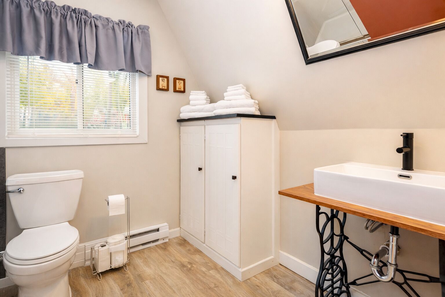 A bathroom with a toilet, a sink on a wooden shelf, a cabinet, and a window with blinds.
