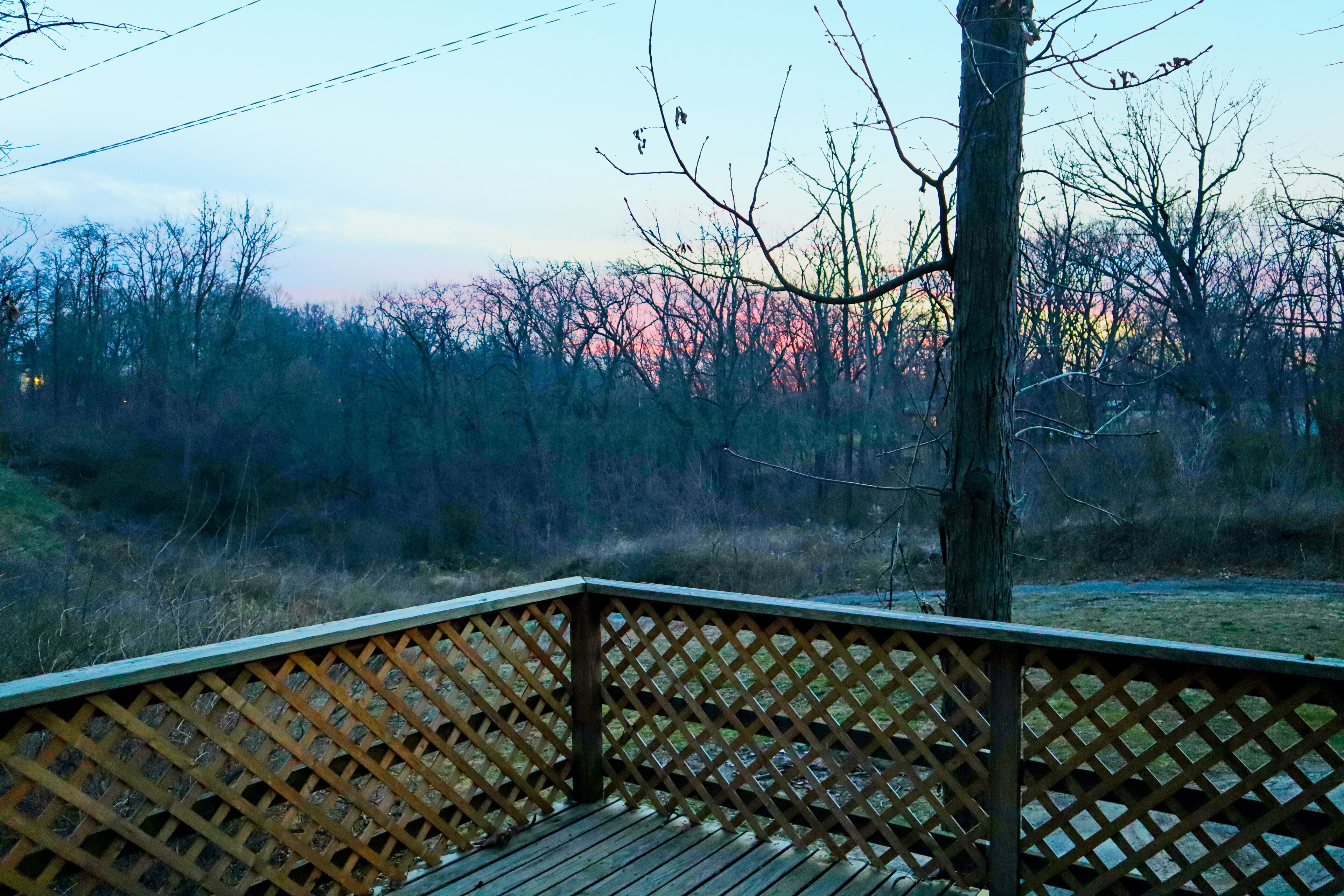 Una terraza de madera con barandilla de celosía que da a un paisaje natural con árboles y una puesta de sol al fondo.