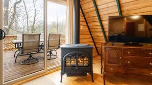 Un acogedor interior de cabaña con estufa de leña, un televisor sobre un mueble de madera y una terraza con mobiliario exterior.