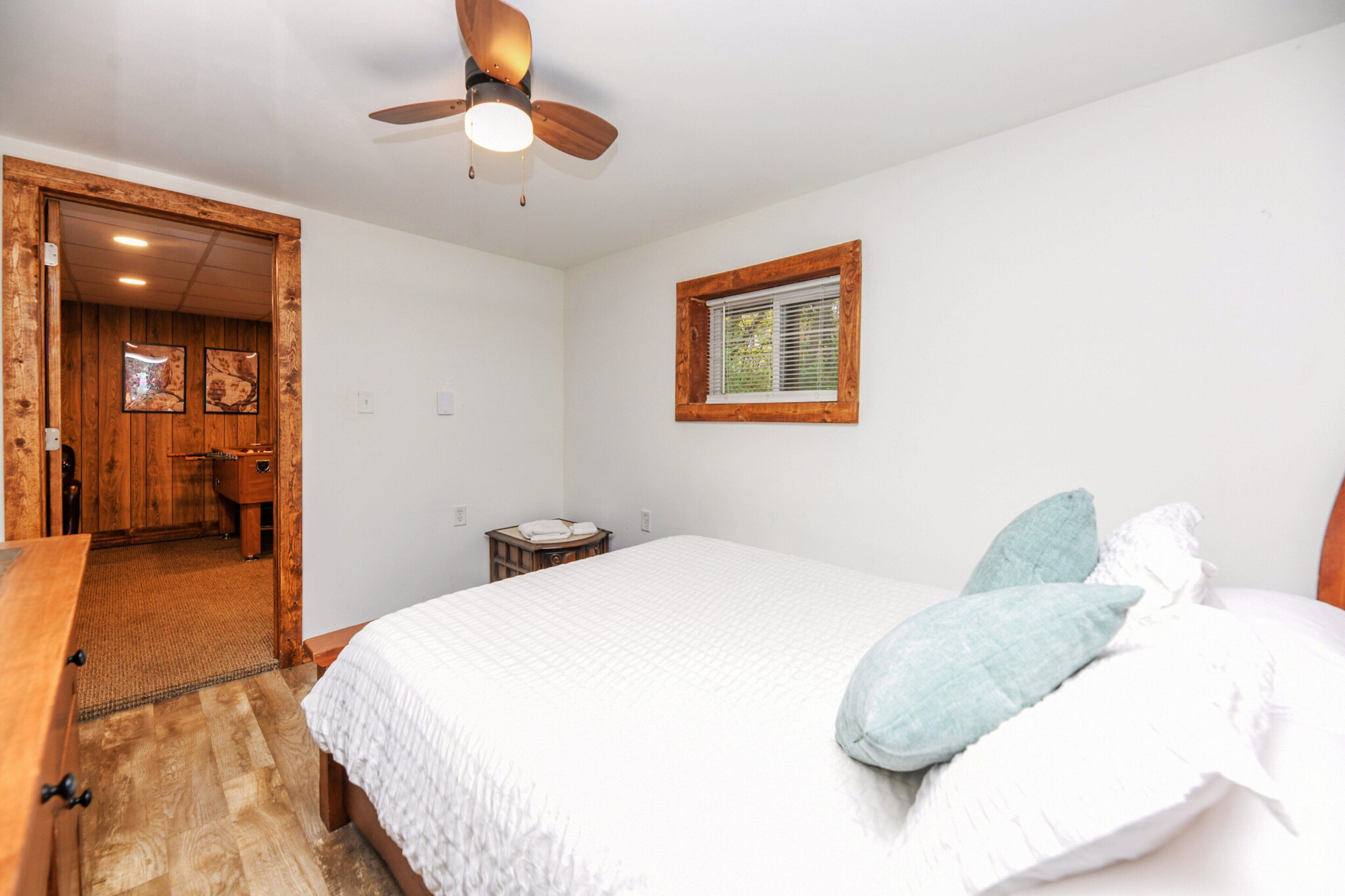 Une chambre avec une porte en bois, un ventilateur de plafond, une fenêtre, un lit avec une literie blanche et une petite table de chevet.