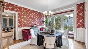 Comedor con una mesa lista para cuatro, rodeado de un papel tapiz floral, una araña y vistas a una casa a través de amplios ventanales.