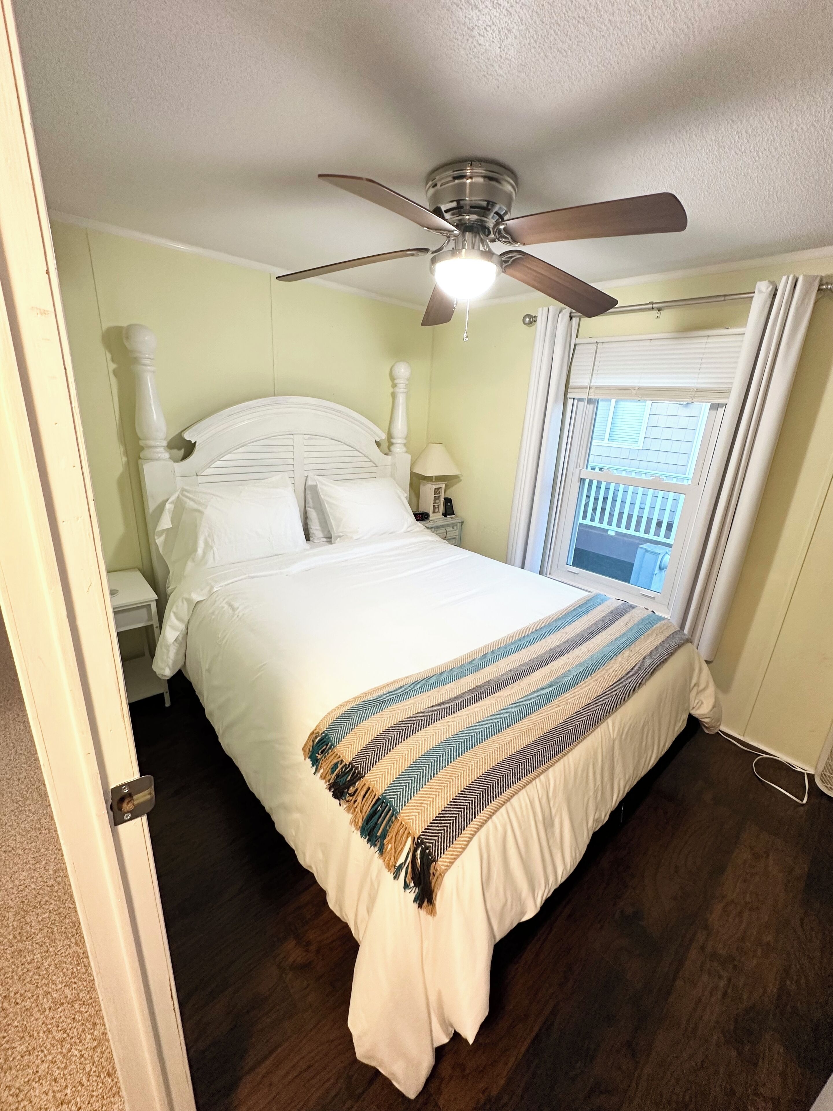 A bedroom with a bed, a ceiling fan, a nightstand, a window with curtains, and a view of a balcony.