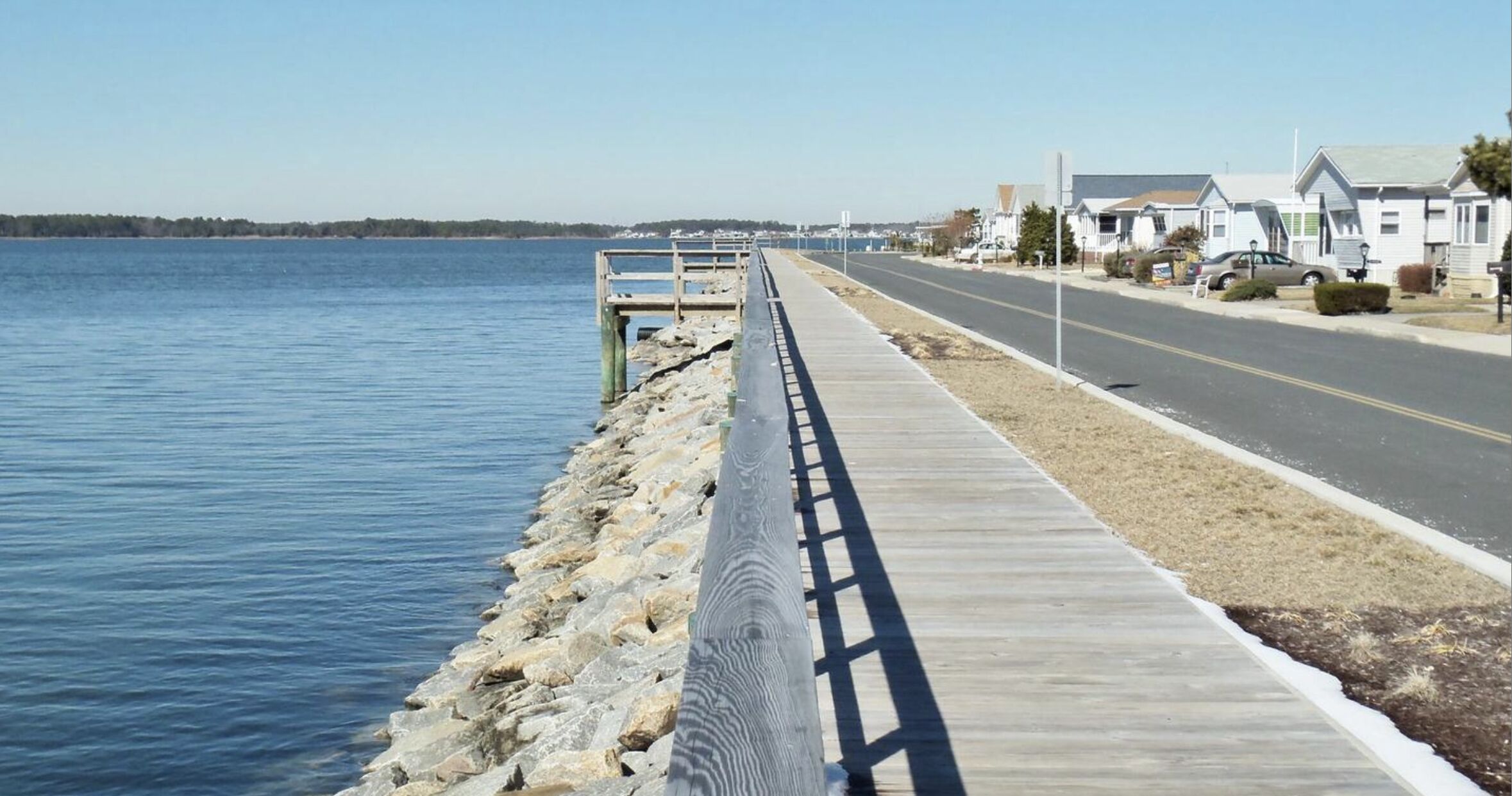 Een kustweg met een houten pier die het water in reikt, omzoomd met huizen en een heldere lucht.