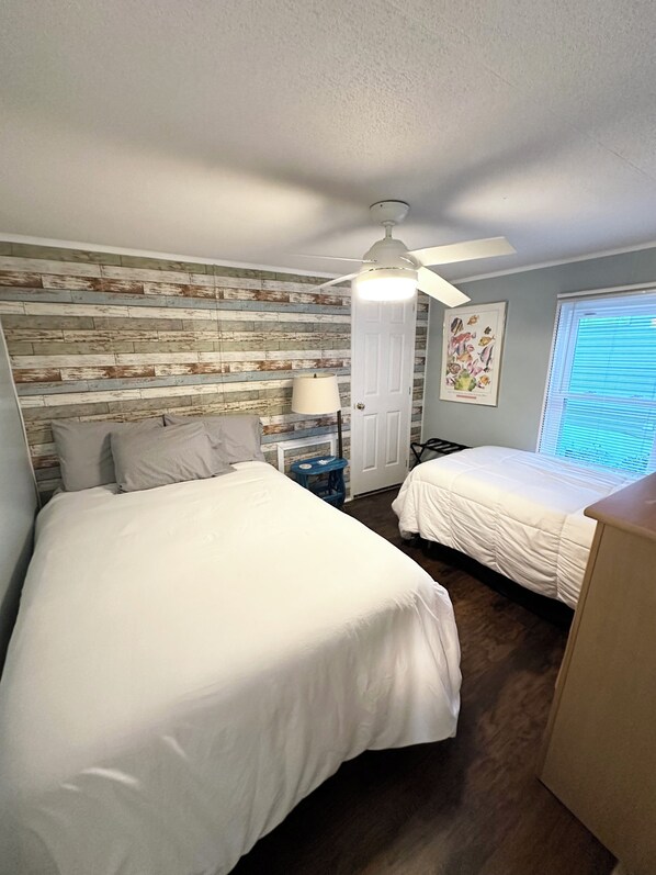 A bedroom with two beds, a ceiling fan, a window with blinds, and a wall with a rustic wood panel design.