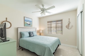 Un dormitorio con cama, ventilador de techo, ventana con persianas, televisor montado en la pared y un adorno decorativo en la pared.