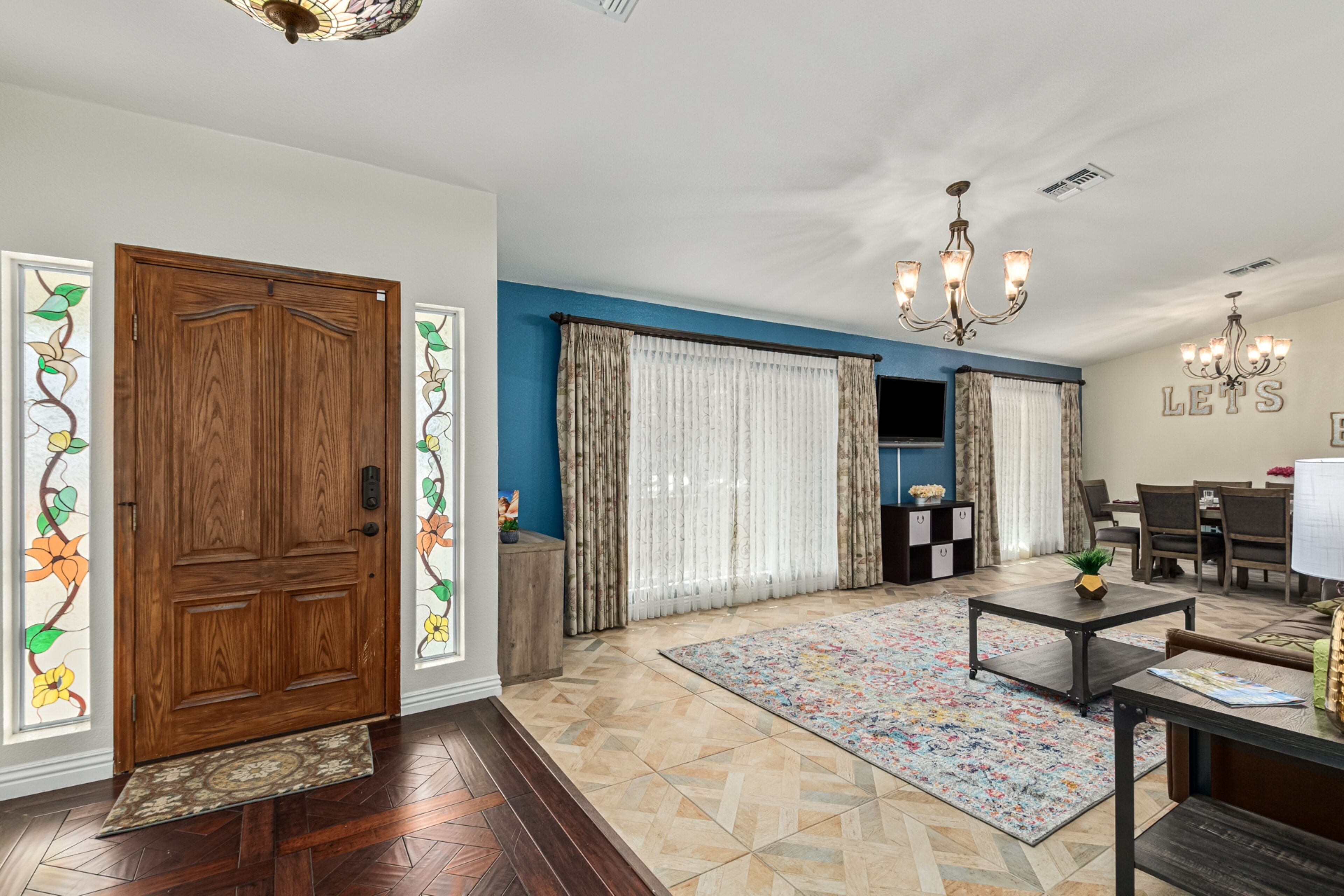 A spacious living room with a wooden door, blue walls, a patterned rug, and a dining area with a chandelier.