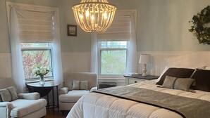 A bedroom with a large bed, two bedside tables, a chandelier, and a window with curtains.
