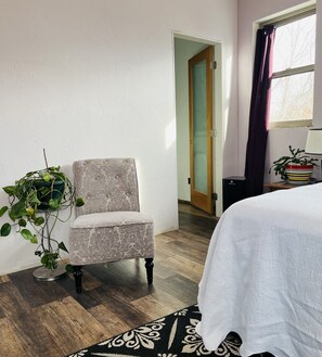 Une pièce comprenant un fauteuil à motifs, une table de chevet avec une lampe, une plante en pot et un lit recouvert d’une courtepointe blanche.
