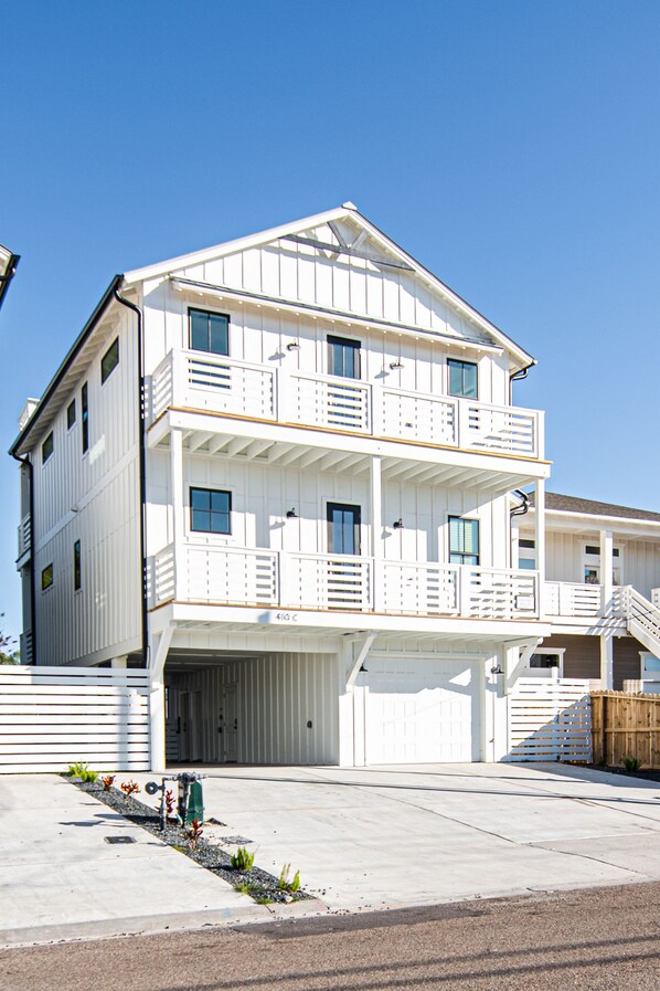 A two-story white house with a garage, a balcony on the second floor, and a wooden fence in the background. - Old Town Road House- 5/Br/4 Ba-Private Heated Pool-Ocean/Bay Views-Rooftop Deck (Port Aransas)