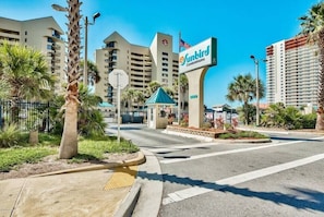 Vista de la calle de un complejo de condominios, con un letrero llamativo que dice 'Sunbird Condominiums', una señal de stop y una pequeña construcción con techo azul.