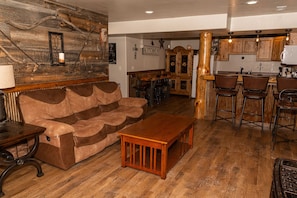 A cozy living room with a brown sofa, wooden coffee table, and a rustic wall. A kitchen area with wooden cabinets and bar stools is visible in the background. - Muley Hill Lodge-Weddings, Events, Rally, Campsites, and Large Parking Lot (Sturgis)