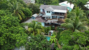 Ein zweistöckiges Haus, umgeben von dichter tropischer Vegetation, darunter Palmen und verschiedene Sträucher.