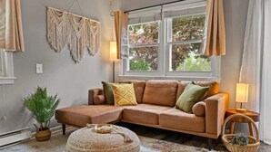 A cozy living room with a brown leather sofa, a round ottoman, a macrame wall hanging, and a window with curtains.