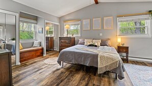 A bedroom with a large bed, a ceiling fan, a dresser, a bench, and a window with a view of trees.