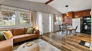 A spacious living room with a brown leather sofa, a dining table with chairs, and a kitchen area with wooden cabinets and a refrigerator.