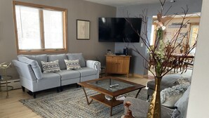 Un salon avec un canapé gris, une table basse, un téléviseur et, au fond, un coin repas.