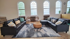A spacious living room with a black leather sectional sofa, a woven ottoman, and a patterned rug. There are three arched windows providing natural light, and a radiator under the windows. - McKinley Mansion Full Building w/Bball gym, Arcades, 13000 sqft, Sleeps 70.  (Pocatello)