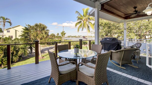 A covered outdoor area with a round glass table and wicker chairs, overlooking a landscaped garden with palm trees and a view of a building across the water. - Winter Getaway by the Bay. Direct Water View! Near Marco Island Beaches. (Naples)