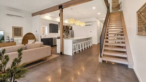 A modern living room with a sofa, a dining area with stools, a kitchen, and a wooden staircase.
