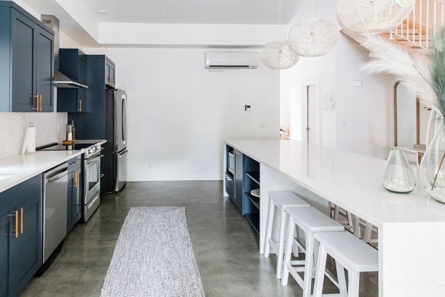 Una cocina moderna con armarios azul oscuro, electrodomésticos de acero inoxidable y encimera blanca. Hay taburetes de bar blancos y una alfombra blanca en el suelo.