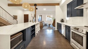 A modern kitchen with dark cabinets, a white countertop, and stainless steel appliances. There is a wooden staircase on the left and a dining area in the background.