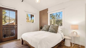A bedroom with a bed, two windows, a wooden floor, a door, and a small bedside table with a lamp.
