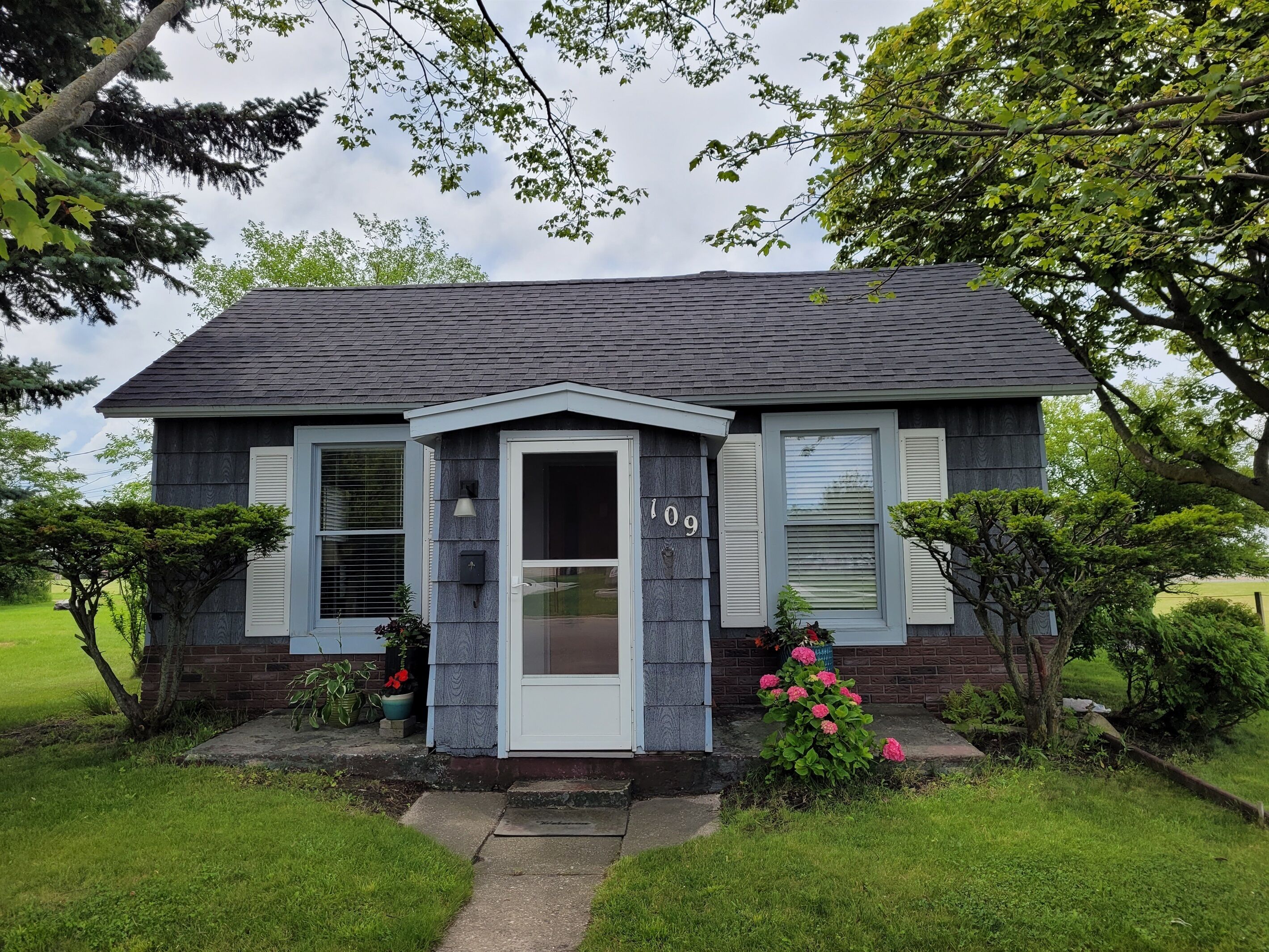A two-story house with a dark gray roof and light gray siding, number 109, surrounded by lush greenery and a well-maintained lawn.