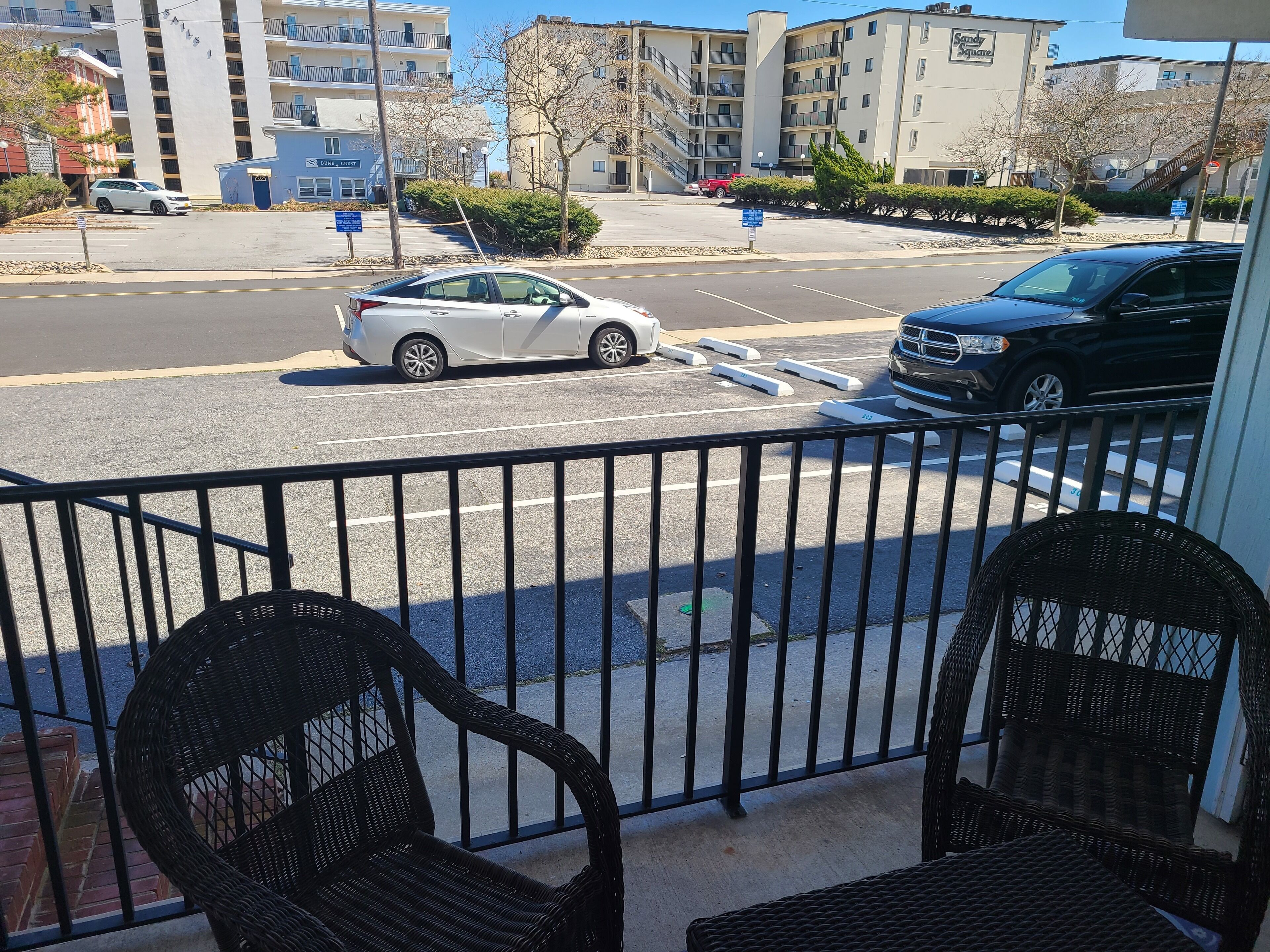 Un balcon avec des fauteuils en osier donnant sur une rue bordée de voitures garées, avec des bâtiments en arrière-plan.