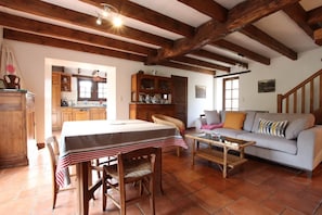Un intérieur rustique avec des poutres apparentes, un coin repas avec une table et des chaises, un canapé et, au fond, une cuisine.
