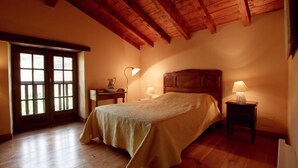 Un dormitorio con techo de madera, una cama con colcha color crema, dos mesitas de noche con lámparas y una ventana con vistas al exterior.