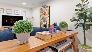 Ein modernes Wohnzimmer mit einem Holztisch, zwei Blumentöpfen, einem farbenfrohen Gemälde und einem Fernseher an der Wand.