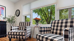 Ein Zimmer mit einem karierten Sessel und einer Couch, einem kleinen Tisch mit Blumen in einer Vase, einer Uhr an der Wand und einem Fenster mit Blick auf Bäume.