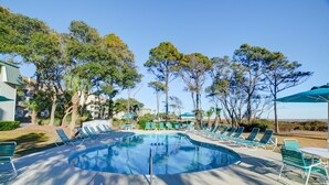 A swimming pool with lounge chairs and umbrellas, surrounded by trees and buildings. - Totally remodeled Oceanfront Villa! New Owner! (Hilton Head Island)