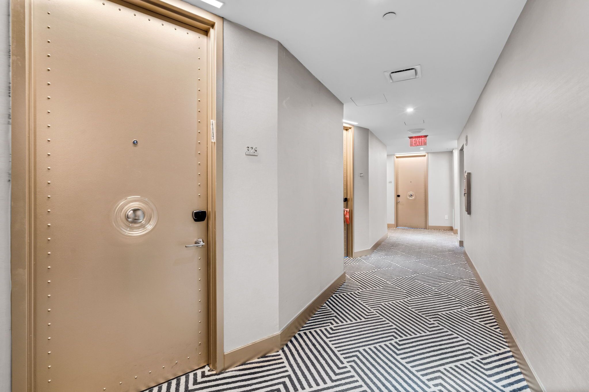A hallway with patterned carpet, beige doors, and a red exit sign.