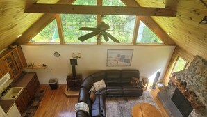 A cozy loft-style room with a wooden ceiling, a large window, a fireplace, a black leather sofa, and a stone fireplace surround.