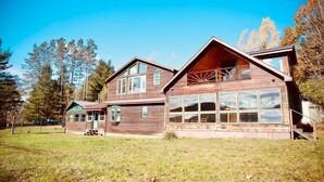 A two-story wooden cabin with a balcony, surrounded by a grassy area and trees.