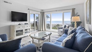 Una sala de estar moderna con un televisor de pantalla plana grande, un mueble blanco, una mesa de centro de vidrio y un sofá azul.