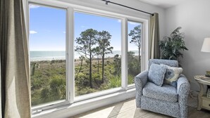 Una habitación con un ventanal grande que da a una playa y un bosque, un sillón estampado y una mesita con una lámpara.