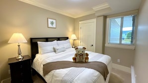 A neatly made bed with a striped comforter, two bedside lamps, a teddy bear, a framed picture on the wall, and a window with blinds. - New two-bedroom legal suite with parking (White Rock)