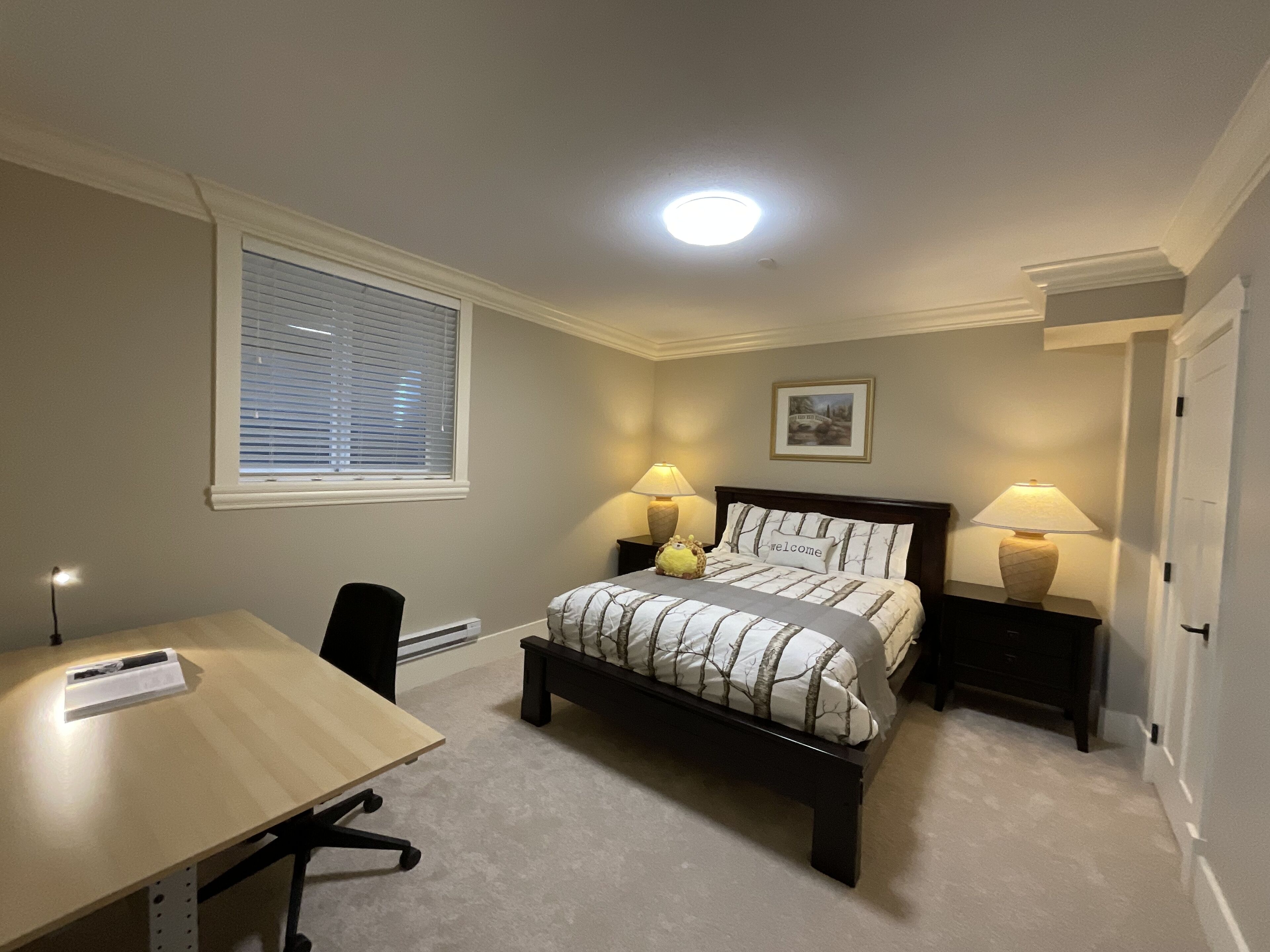 Une chambre à coucher comprenant un lit, deux tables de chevet avec des lampes, un bureau, une chaise, une fenêtre avec des stores et un luminaire au plafond.