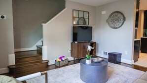 A modern living room with a staircase, a flat-screen TV, a decorative plate, and a small plant on a round table.