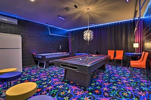 Una mesa de billar con alfombra estampada en motivos de estrella, un refrigerador y sillas naranjas en una habitación con iluminación LED azul.