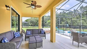 A covered outdoor seating area with wicker furniture and a ceiling fan, overlooking a swimming pool and greenery. - Private Pool! Game room! Minutes to Disney! Themed rooms! (Kissimmee)