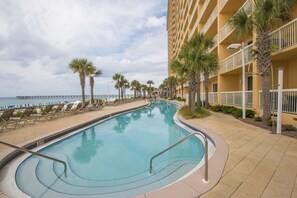 A swimming pool with a curved design, surrounded by lounge chairs and palm trees, adjacent to a hotel building with balconies.