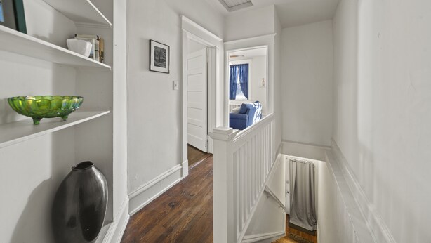 A hallway with white walls, wooden flooring, and a staircase with a white railing. There are shelves with various items and a door leading to another room. - Walk Central. Museum district. Scott's Addition. 2BR - King and full bed.  (Richmond)