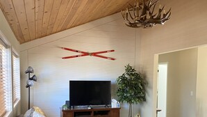 Una sala de estar con techo de madera, un televisor sobre un mueble de madera, un sofá con cojines, una mesa de centro y una alfombra estampada.